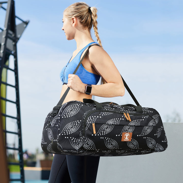 Fit woman with a ponytail wearing a blue sports bra, carrying a black and white paisley duffle bag with yoga mat holder over her shoulder, walking outdoors, blue sky