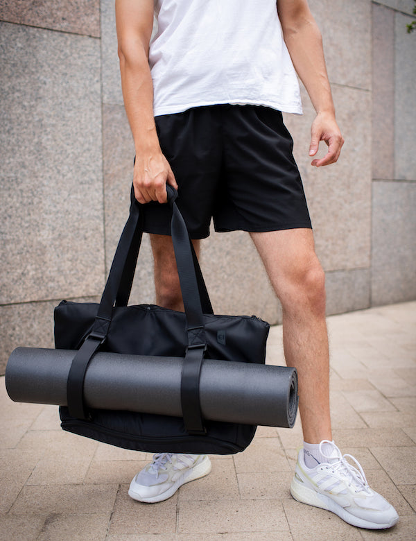 Man wearing a white singlet carrying Warrior2 large black yoga mat tote with front straps, over the shoulder, standing against a stone wall.