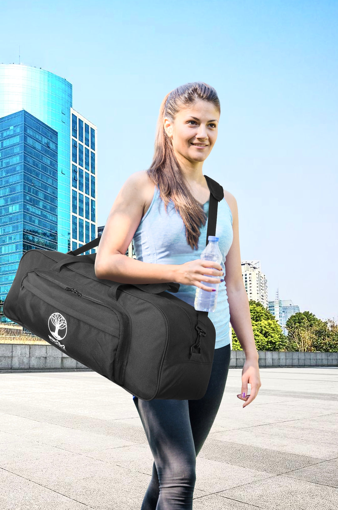 Woman carrying a Warrior2 womens gym bag with yoga mat holder while holding a water bottle outdoors with city buildings in the background, blue sky.