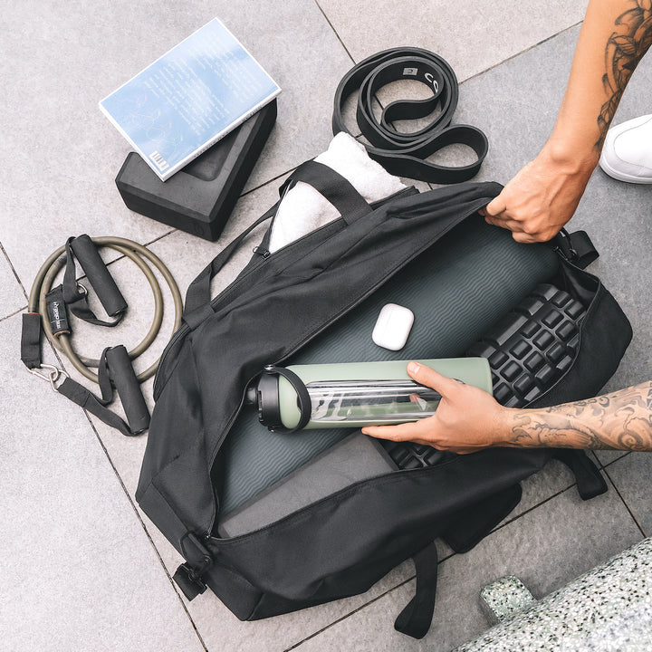 Person organizing gym equipment into a warrior2 black yoga gym bag on a tiled floor.
