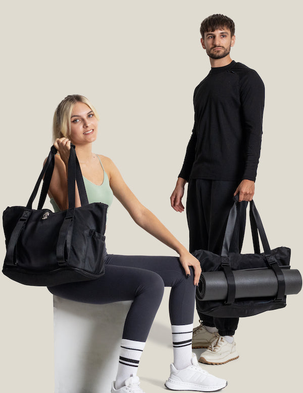 Woman sitting and man standing, both holding black Warrior2 yoga gym totes; the man’s bag includes a rolled yoga mat in front straps.
