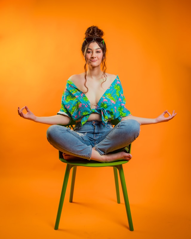 woman in a colorful top sits cross-legged in a chair meditating against an orange backdrop, showcasing lifestyle with yoga backpacks