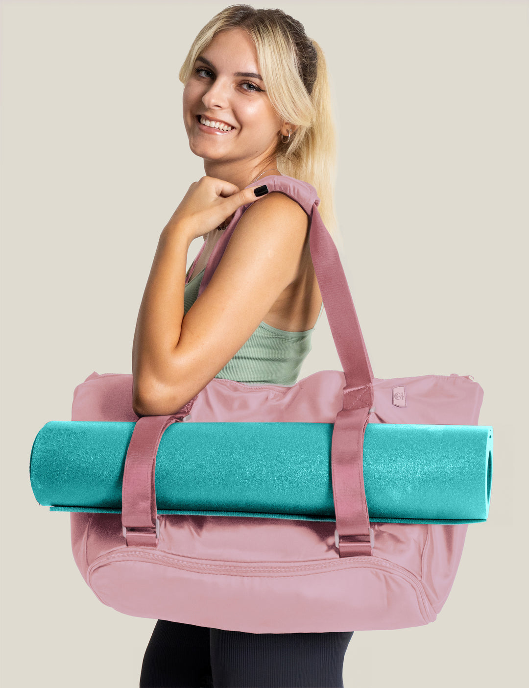 blonde Woman smiling at the camera holding a warrior2 pink yoga duffel tote bag with a teal yoga mat, smiling against a plain background